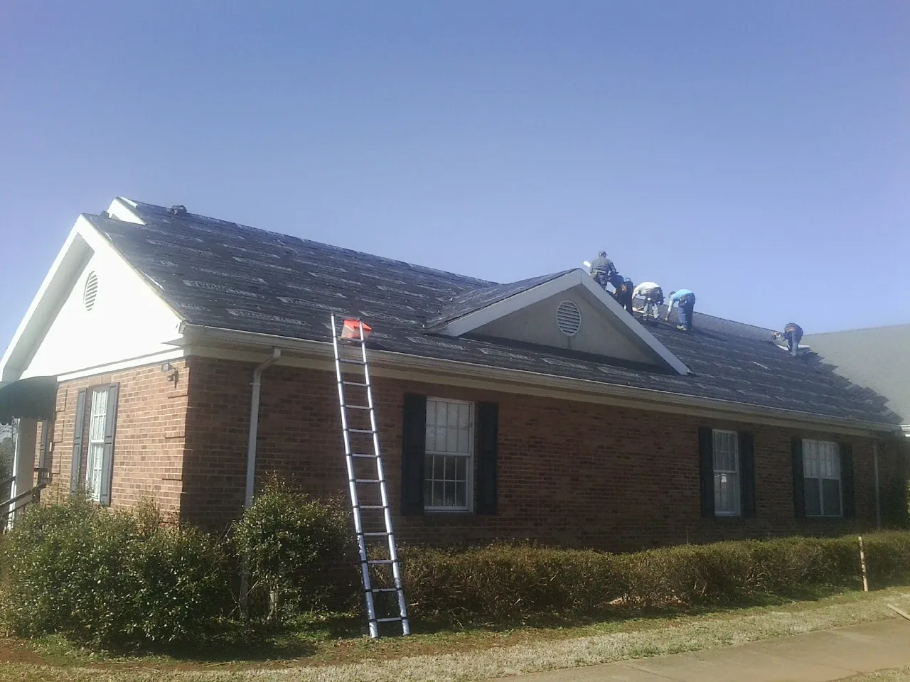 Crew tearing shingles off a brick ranch roof with a ladder set — Emergency Roof Repair work by our crew in Ramah