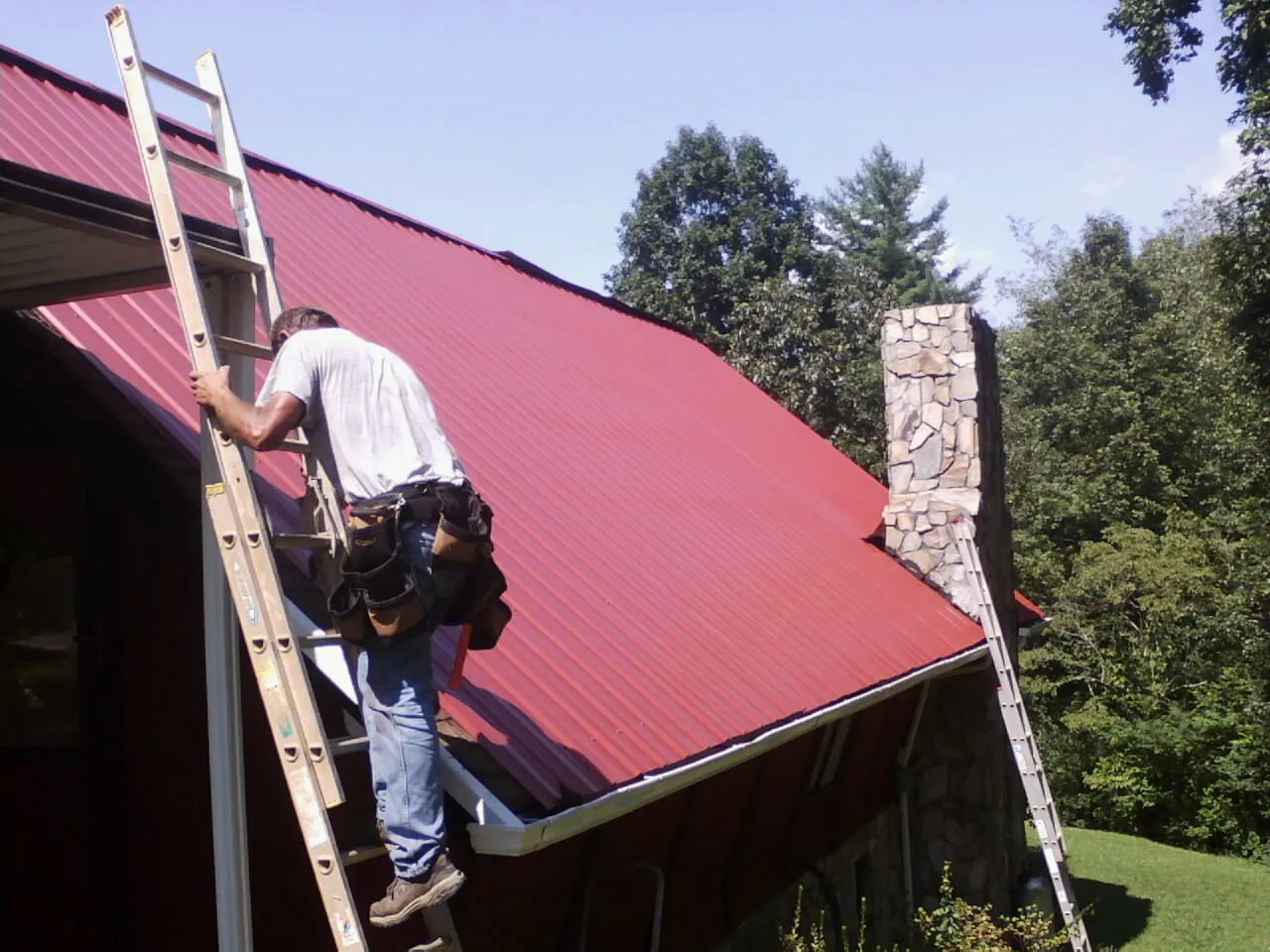 Roofer on a ladder beside a red metal roof and stone chimney — Soffit & Fascia Repair by Roofing Brothers in Ramah